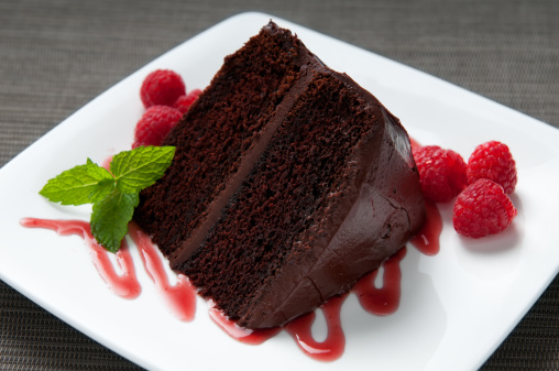 Close up and selective focus on a slice of a layered chocolate cake with icing surrounded by fresh raspberries on a white plate decorated with raspberry sauce and mint.