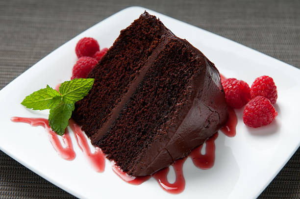 Layered Chocolate Cake with Frosting, Raspberries, Sauce and Mint Close up and selective focus on a slice of a layered chocolate cake with icing surrounded by fresh raspberries on a white plate decorated with raspberry sauce and mint. chocolate cake stock pictures, royalty-free photos & images