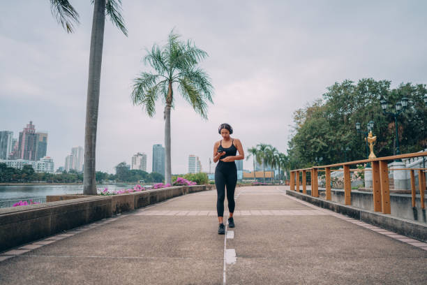 Female athlete using phone in a city park Young woman is using phone while relaxing from jogging in the urban park with modern city skyline. Walking stock pictures, royalty-free photos & images
