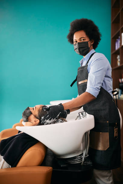 Black hairdresser wearing face mask while washing customer's hair at the salon during COVID-19 pandemic. African American hairstylist washing woman's hair at the salon and looking at camera. They are wearing face masks due to coronavirus pandemic. black woman washing hair stock pictures, royalty-free photos & images