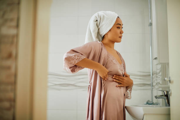 Asian woman doing breast self-exam while looking herself in a mirror in the bathroom.