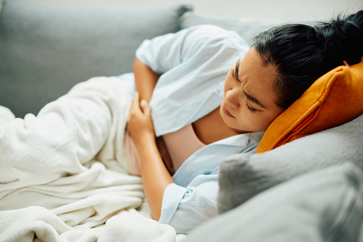 Young Asian woman lying down on sofa while experiencing stomach pain.