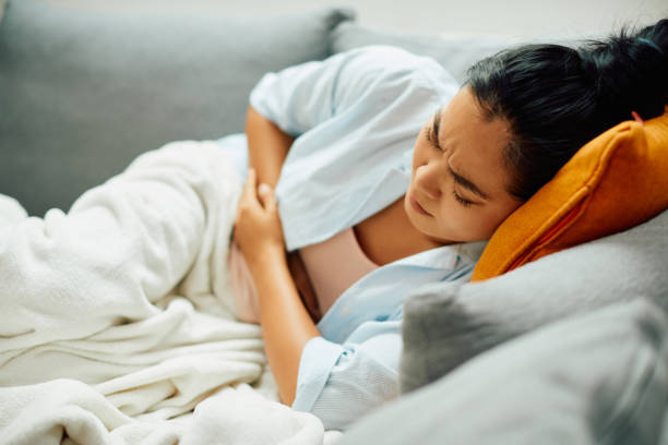 Young Asian woman having stomachache while lying down on sofa at home. Young Asian woman lying down on sofa while experiencing stomach pain. food poisoning stock pictures, royalty-free photos & images