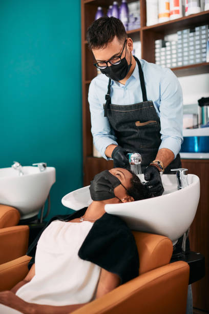 Hairdresser wearing face mask while washing black woman's hair at the salon. African American woman and her hairstylist wearing protective face mask during hair wash at the salon. black woman washing hair stock pictures, royalty-free photos & images