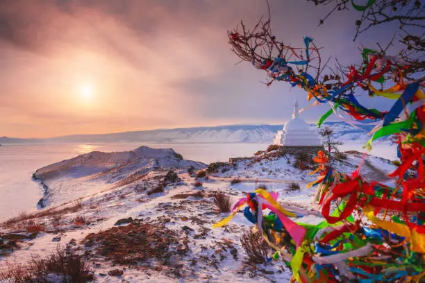 Buddhist stupa and tree with colorful prayer ribbons on Ogoy island on Baikal lake. Baikal, Siberia, Russia. Winter landscape at sunset.