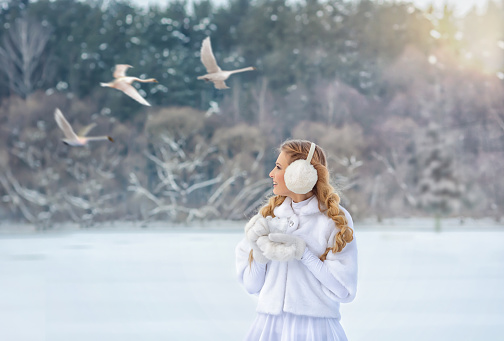 Young blonde woman in white clothes in winter looking at white flying swans in the sky in the background smiling. Swan Lake.