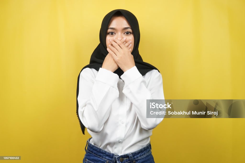Beautiful young asian muslim woman shocked, surprised, disbelieving, getting shocking information, with hands covering mouth isolated on yellow background Fear Stock Photo