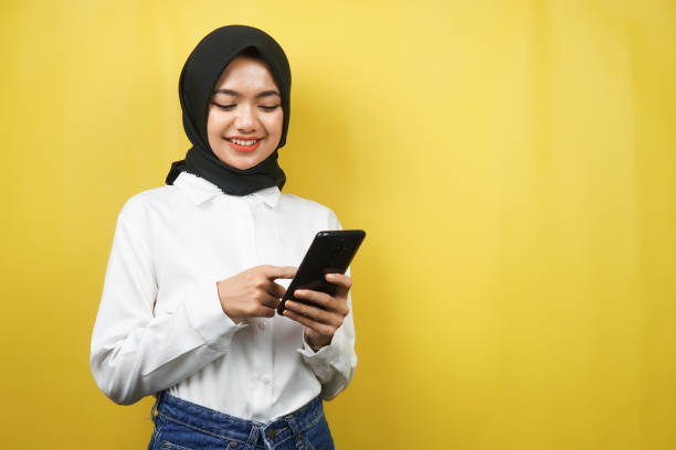 Beautiful young asian muslim woman smiling confidently holding smartphone isolated on yellow background, advertising concept stock photo