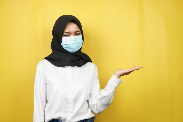 Muslim woman wearing medical mask, hands presenting something in empty space, isolated on yellow background stock photo