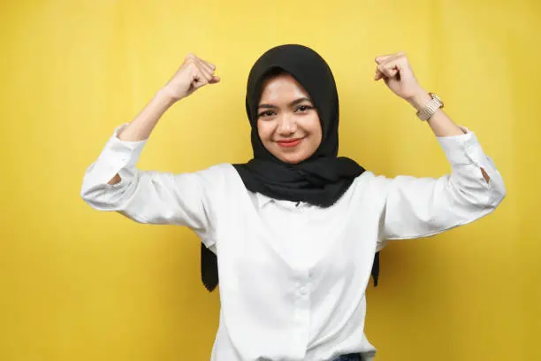 Photo of Beautiful asian young muslim woman with raised muscles, strength sign arms, isolated on yellow background