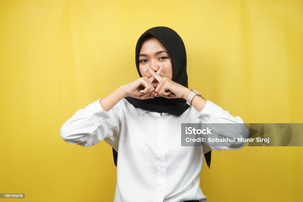 Beautiful asian young muslim woman with finger on mouth, telling to be quiet, don't make noise, lower your voice, don't talk, isolated on yellow background Gossip Stock Photo