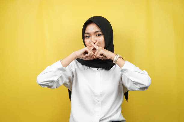 Beautiful asian young muslim woman with finger on mouth, telling to be quiet, don't make noise, lower your voice, don't talk, isolated on yellow background stock photo