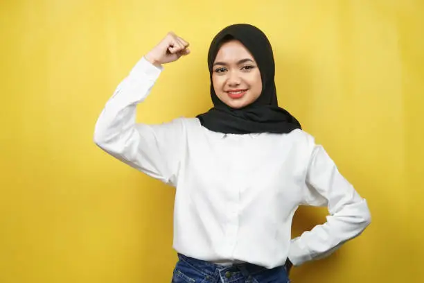 Photo of Beautiful asian young muslim woman with raised muscles, strength sign arms, isolated on yellow background
