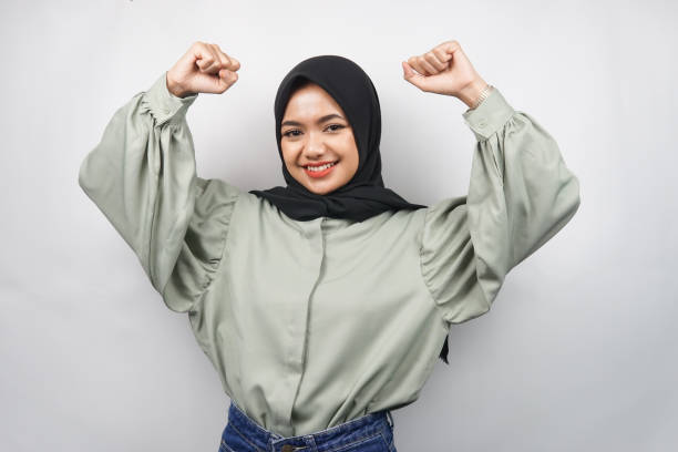 Beautiful young asian muslim woman confident and smiling, with clenched fist, punching, sign for spirit, fighting, victory sign, isolated on gray background stock photo