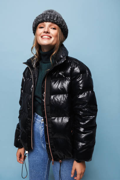 istock Young pretty smiling woman in knitted hat and black down jacket joyfully looking in camera over blue background 1357105549