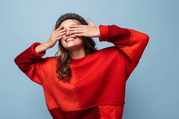 schönes lächelndes mädchen mit welligem haar in hut und rotem pullover, das glücklich die augen mit den händen über blauem hintergrund bedeckt - hands covering eyes stock-fotos und bilder