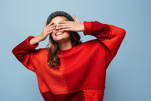 Beautiful smiling girl with wavy hair in hat and red sweater happily covering eyes with hands over blue background