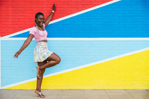 Cheerful african-american woman in colorful skirt against a multicolored wall. Woman with braids standing on one leg and arms outstretched looking to the side. Concept of people.