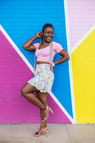 Very happy black African woman posing on a wall in many colors. Lifetyle photo of a happy African woman Cheerful african-american woman in colorful skirt against a multicolored wall. Woman with braids standing on one leg and arms outstretched looking to the side. Concept of people. black hair braiding stock pictures, royalty-free photos & images
