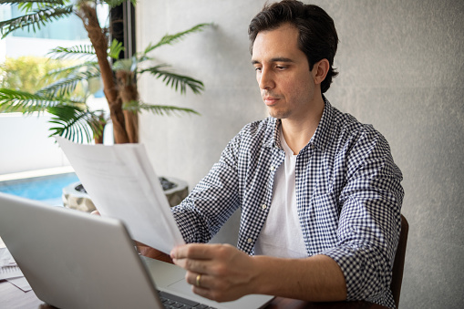 Mid adult man reading a paper and working on the laptop at home
