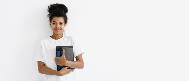 Study, education, advertising concept. Web banner of happy young african american woman standing with books and notepad in hands against white copy space background, smiling wide, looking at camera