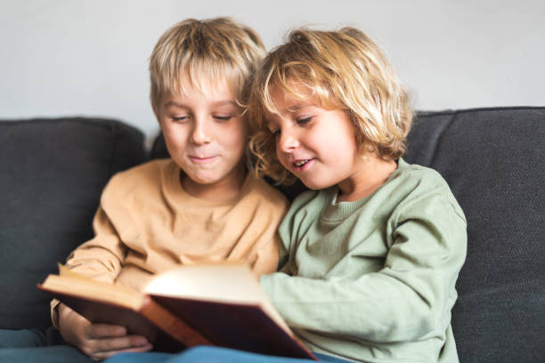 Happy little kids with blond hair and casual wear smiling while reading interesting story sitting together on comfortable couch