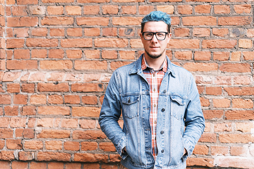 Stylish and fashionable handsome young man with blue hair wearing in jeans jacket standing on background of brick wall. Model looks at camera and smiling.