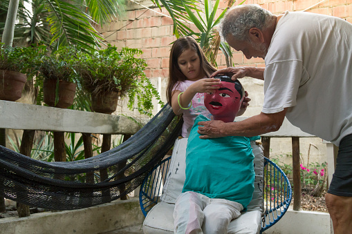 A mature latin man sewing a t-shirt and pants to make a kind of doll / monigote / stuffed dummies / Paper Mache effigy, to burn at midnight of New Year's Eve in Ecuador, while sharing time with his little young latin granddaughter.