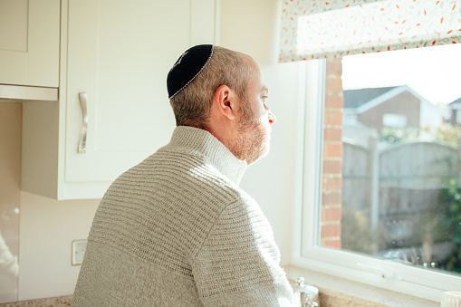 Color image depicting a mid adult Jewish man in his 30s wearing a traditional Jewish skull cap (with star of David design). The man has a beard and is relaxing at home. Room for copy space.
