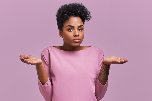 Uncertain African American female with dark skin, shruggs shoulders and asks something, has clueless expression, isolated on lavender background. Woman faces dilemma.