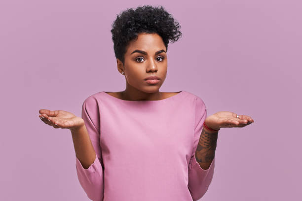 Uncertain African American female with dark skin, shruggs shoulders and asks something, has clueless expression, isolated on lavender background. Woman faces dilemma Uncertain African American female with dark skin, shruggs shoulders and asks something, has clueless expression, isolated on lavender background. Woman faces dilemma. Confusion stock pictures, royalty-free photos & images
