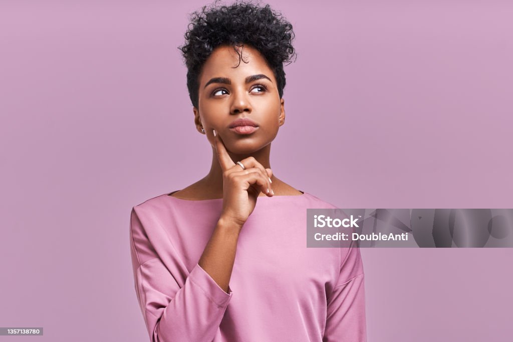Pensive attractive curly African American female being deep in thoughts, raises eye, wears fashionable clothes, stands against lavender wall Pensive attractive curly African American female being deep in thoughts, raises eye, wears fashionable clothes, stands against lavender wall. Uncertainty Stock Photo