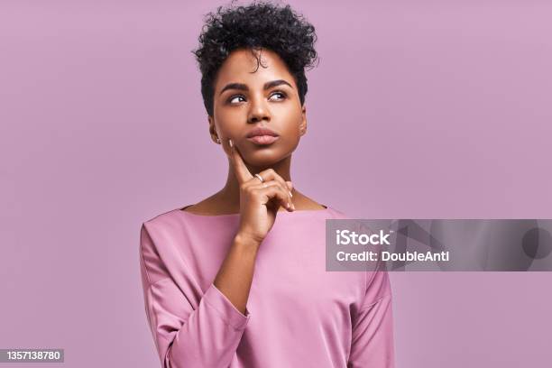 Pensive Attractive Curly African American Female Being Deep In Thoughts Raises Eye Wears Fashionable Clothes Stands Against Lavender Wall Stock Photo - Download Image Now