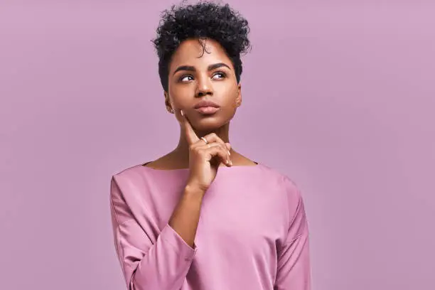 Photo of Pensive attractive curly African American female being deep in thoughts, raises eye, wears fashionable clothes, stands against lavender wall