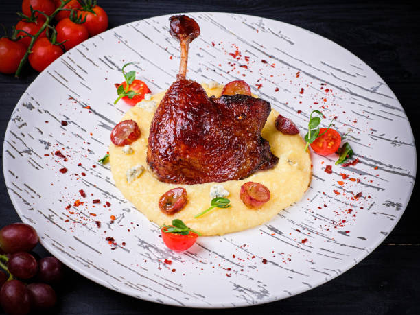 Duck leg confit with mashed potatoes and blue cheese, grapes and tomatoes. Traditional French Duck leg confit served on mashed potatoes with Dor blue cheese, grapes and Cherry tomatoes. Restaurant Main dish on white plate on black wooden background with ingredients confit stock pictures, royalty-free photos & images