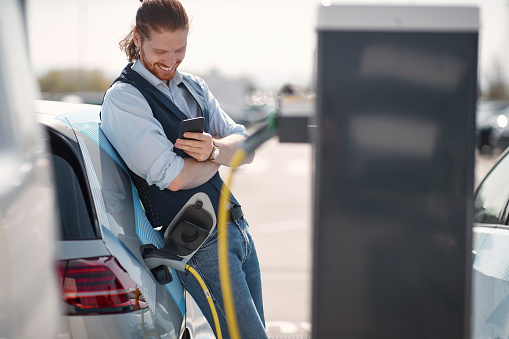 Young smiling man using smartphone near electric car on charging station