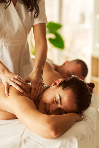 Head and shoulders of satisfied female customer smiling with her eyes closed and head lying on top of her hands while enjoying the benefits of relaxation during a spa massage therapy. Masseuse treats cervical and shoulders muscles.