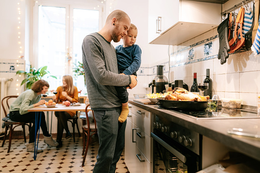 Man carrying his son standing at kitchen counter and looking at Christmas dinner at home. Father and son in kitchen looking at thanksgiving meal on counter.