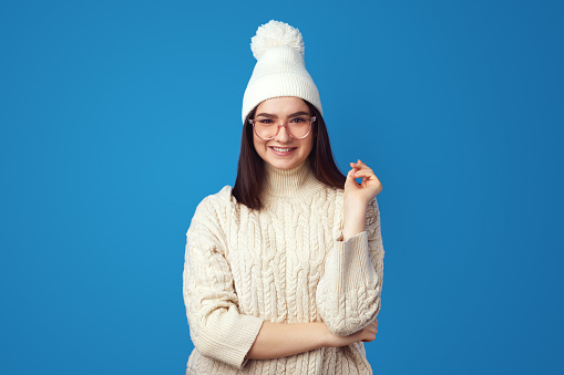 Good looking woman with long hair, wears warm hat and oversized knitted sweater, feels joyful, enjoys winter time, isolated over blue background.