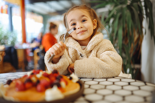 Little girl having crepes for breakfast. The crepes are sprinkled with fruits - blueberries and raspberries. Sweet toddler birthday boy, eating belgian waffle with raspberries and chocolate at cafe