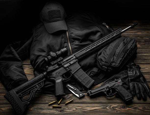 istock Modern automatic rifle with a telescopic sight on a dark background. Jacket, cap, gloves on a wooden table. The uniform of a guard or a mercenary. 1357186588
