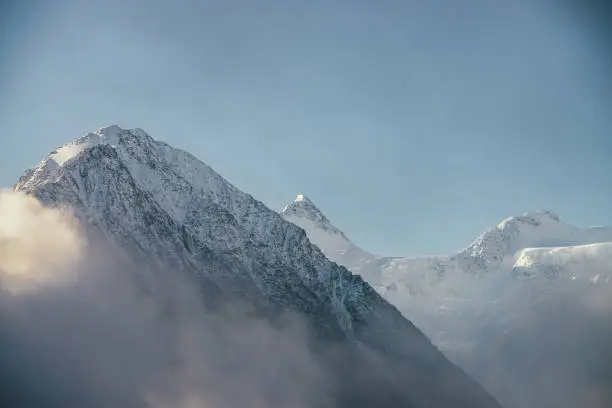 Beautiful view of snow-capped mountains above thick clouds in sunshine. Scenic bright mountain landscape with white-snow peak among dense low clouds in blue sky. Wonderful scenery with snowy pinnacle.