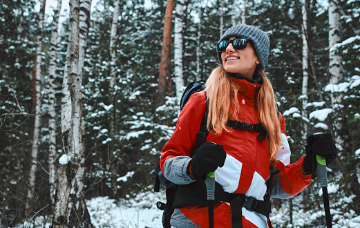 Beautiful young woman dressed in warm sportswear, hat and sunglasses stands with trekking poles in a snowy pine forest. Copy space.