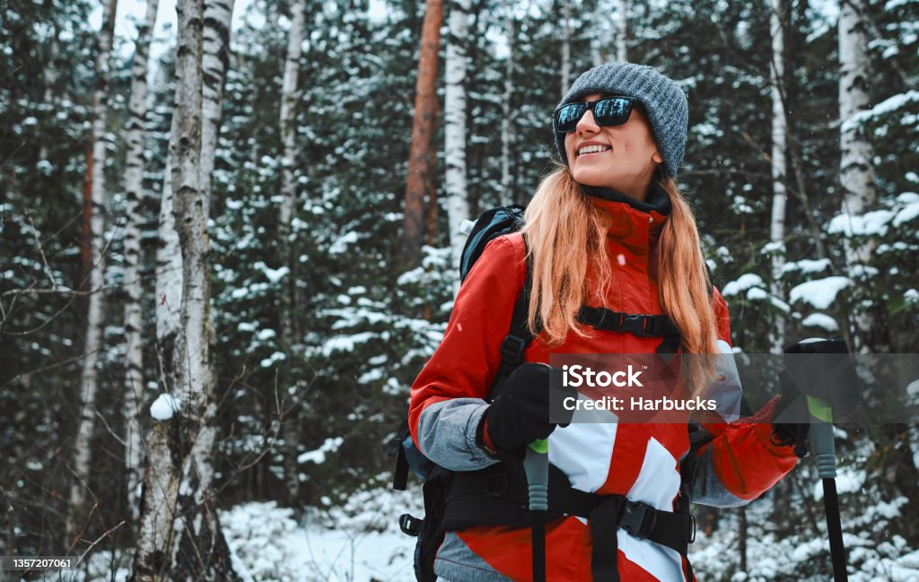 Beautiful young woman dressed in warm sportswear, hat and sunglasses stands with trekking poles in a snowy pine forest. Copy space. Skiing Stock Photo