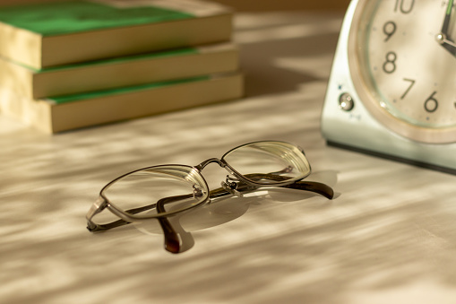Eyeglasses, table clock and books on the desk by the window in the evening