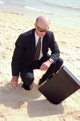 A Businessman at the beach on a sunny day looking at his briefcase stuck in the sand