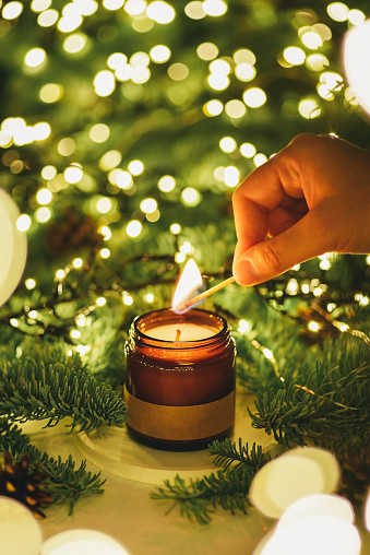 Hand lights a scent candle with a match against the background of green Christmas tree branches and lights. Home comfort concept with aroma.