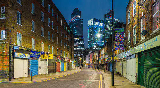 The futuristic towers of the City financial district illuminated at night overlooking the quiet streets and shops of Petticoat Lane Market in Spitafields, London.