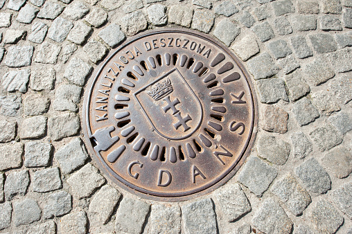 Ornate manhole cover and cobbles, Gdansk, Poland.  Situated on the Baltic coast of Poland Gdańsk (Danzig in German) is the port city, at the centre of which is its ancient old Main Town, reconstructed after WWII, where the colorful facades of Piwna, Long Market, are home to shops and restaurants.