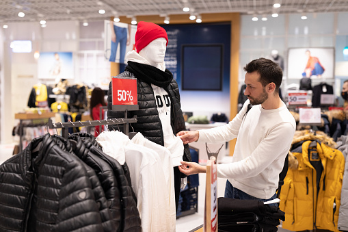 Adult handsome brunet man in white sweatshirt chooses stylish fashionable clothes in shopping mall. Store.